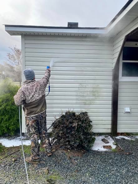The owner in a camo jacket and beanie spraying the vinyl siding of a house with a pressure washing wand, with a visible line of grime still to be cleaned beside him.