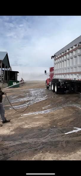 A worker hosing down a concrete commercial yard beside a parked semi trailer.