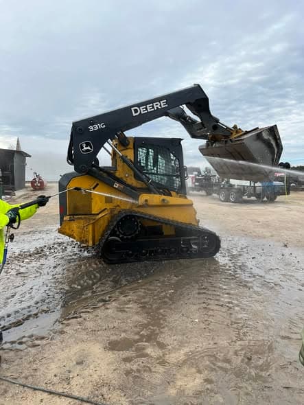 The owner in a hi-vis jacket washing a tracked John Deere skid steer with a pressure washing wand in a working yard.