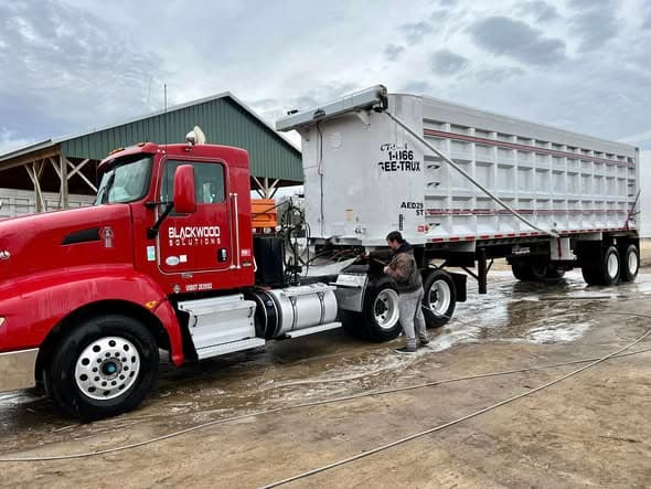 The owner washing the side of a red semi tractor and its dump trailer at a commercial yard.
